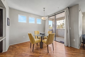Un coin repas avec une table ronde et quatre chaises, un luminaire au plafond, et une vue sur un balcon extérieur accessible par une porte-fenêtre coulissante.