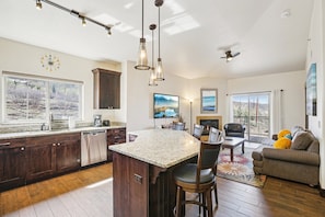 A modern kitchen with a central island, bar stools, a dining area, and a living room with a sofa and a fireplace.