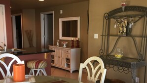 A dining area with a glass table, white chairs, a candle, and a decorative metal shelf with glasses. - Gulf Front Condo, Corner Unit, Stunning Views, Great Restaurants (Panama City Beach)