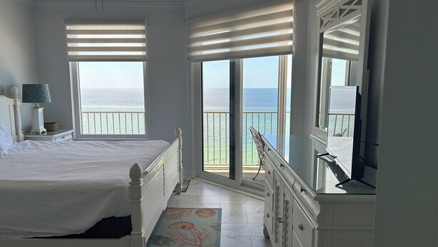 A bedroom with a bed, a dresser, a lamp, and a view of the ocean through large windows. - Gulf Front Condo, Corner Unit, Stunning Views, Great Restaurants (Panama City Beach)