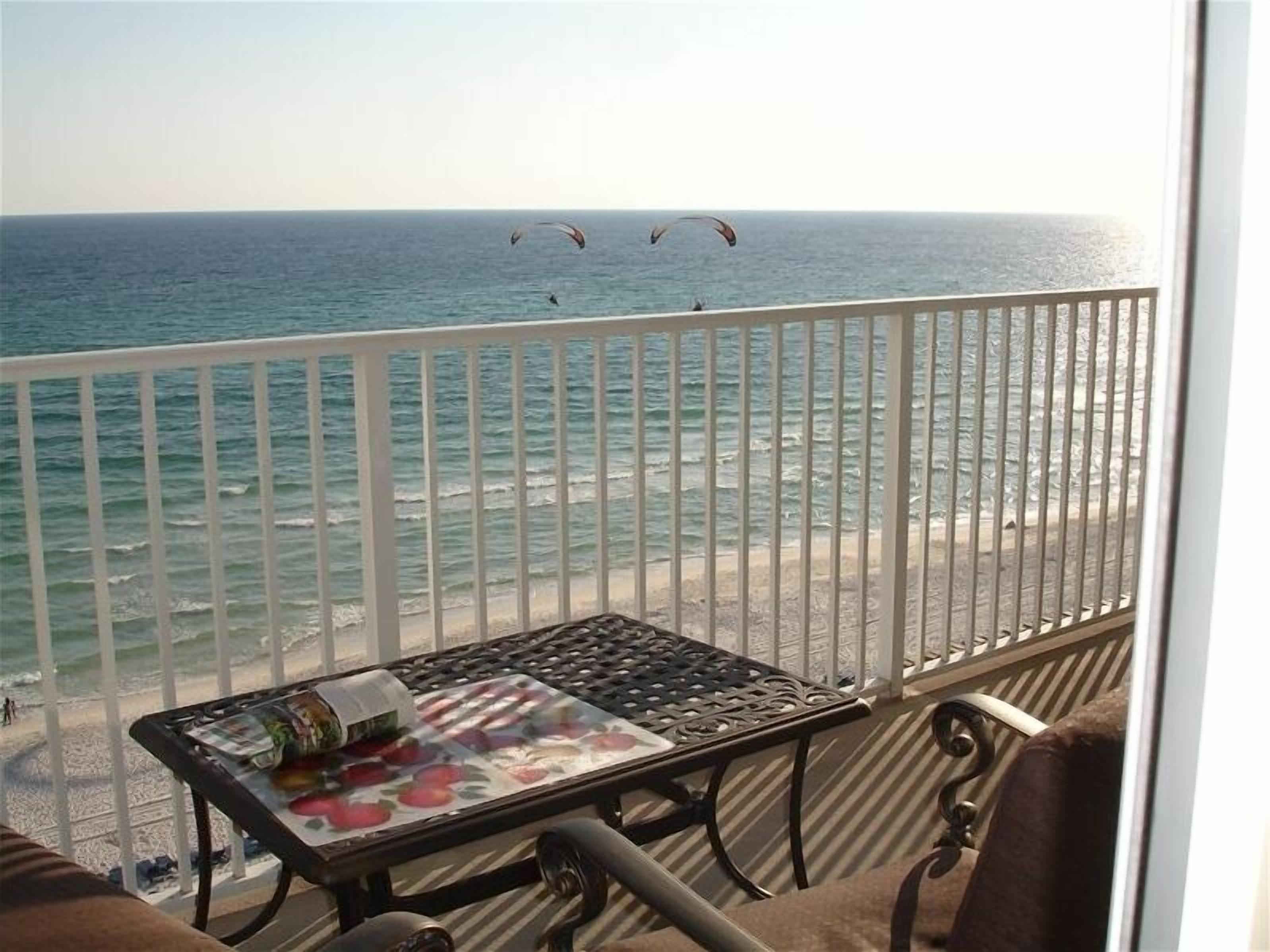 A balcony with a view of the ocean and beach, a table with a fruit basket, and a chair.