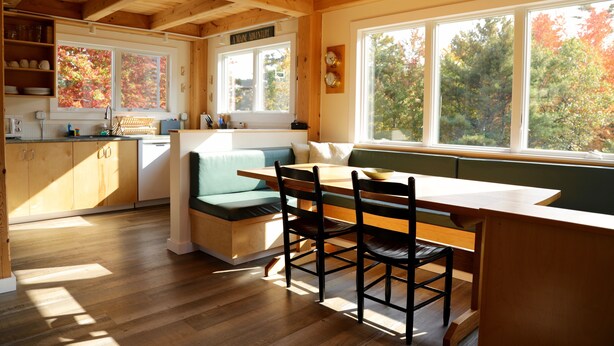 A wooden cabin interior with a dining area, kitchen, and large windows.