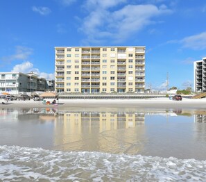 Un hotel frente a la playa con varios balcones, una playa arenosa en primer plano y cielos despejados.