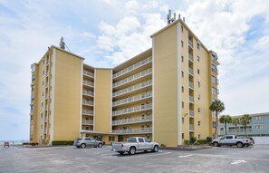 A multi-story yellow building with balconies, a parking lot with several vehicles, and palm trees in the background.