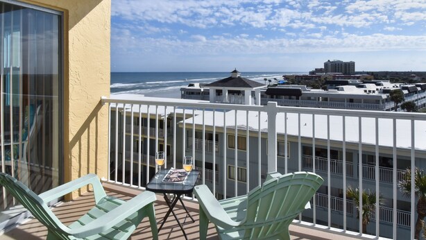 Un balcon avec deux chaises vertes et une petite table, d’où l’on a vue sur une plage et des immeubles.