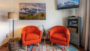 A living room with two orange armchairs, a colorful rug, a lamp, a TV, and a wall-mounted painting. - Cozy 2BR/2BA Condo at Big Sky Resort (Big Sky)