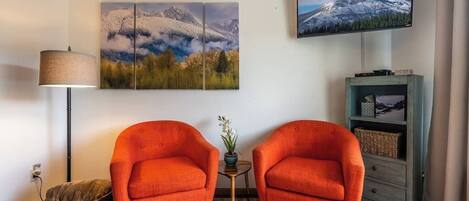Een woonkamer met twee oranje fauteuils, een kleurrijk vloerkleed, een lamp, een televisie en een aan de muur hangend schilderij.