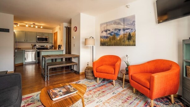 A modern living room with a wooden coffee table, two orange armchairs, a patterned rug, and a painting of a mountain landscape. - Cozy 2BR/2BA Condo at Big Sky Resort (Big Sky)