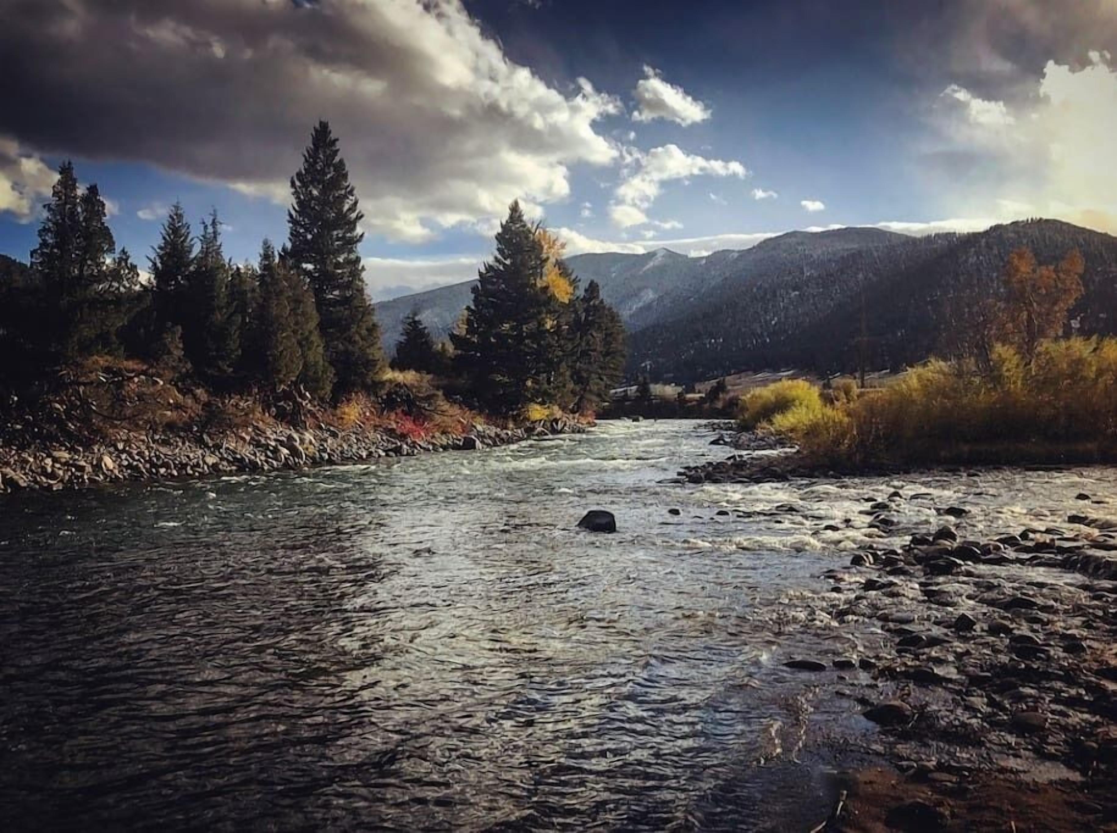 Une rivière à lit rocheux, bordée d’arbres et entourée de montagnes, sous un ciel partiellement nuageux.