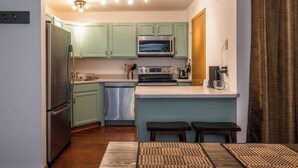 A compact kitchen with light green cabinets, a stainless steel refrigerator, a built-in oven, and a dishwasher. There are two stools at the island and a small countertop with a coffee maker. - Cozy 2BR/2BA Condo at Big Sky Resort (Big Sky)