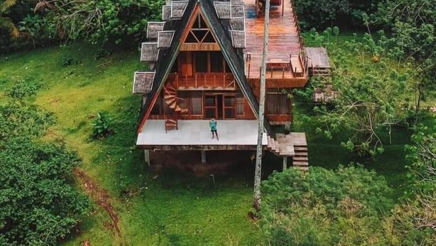 A triangular wooden house surrounded by lush greenery and a view of the ocean.