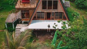 A unique, triangular-shaped wooden house with a thatched roof, surrounded by lush greenery.