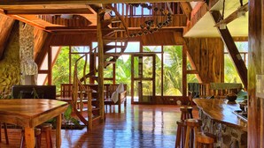 A rustic wooden interior with a spiral staircase, wooden furniture, and large windows offering a view of lush greenery.