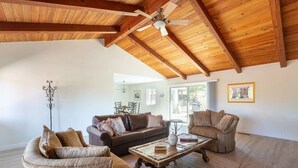 A spacious living room with wooden beams, a ceiling fan, a brown leather sofa, a wicker armchair, and a rustic coffee table.