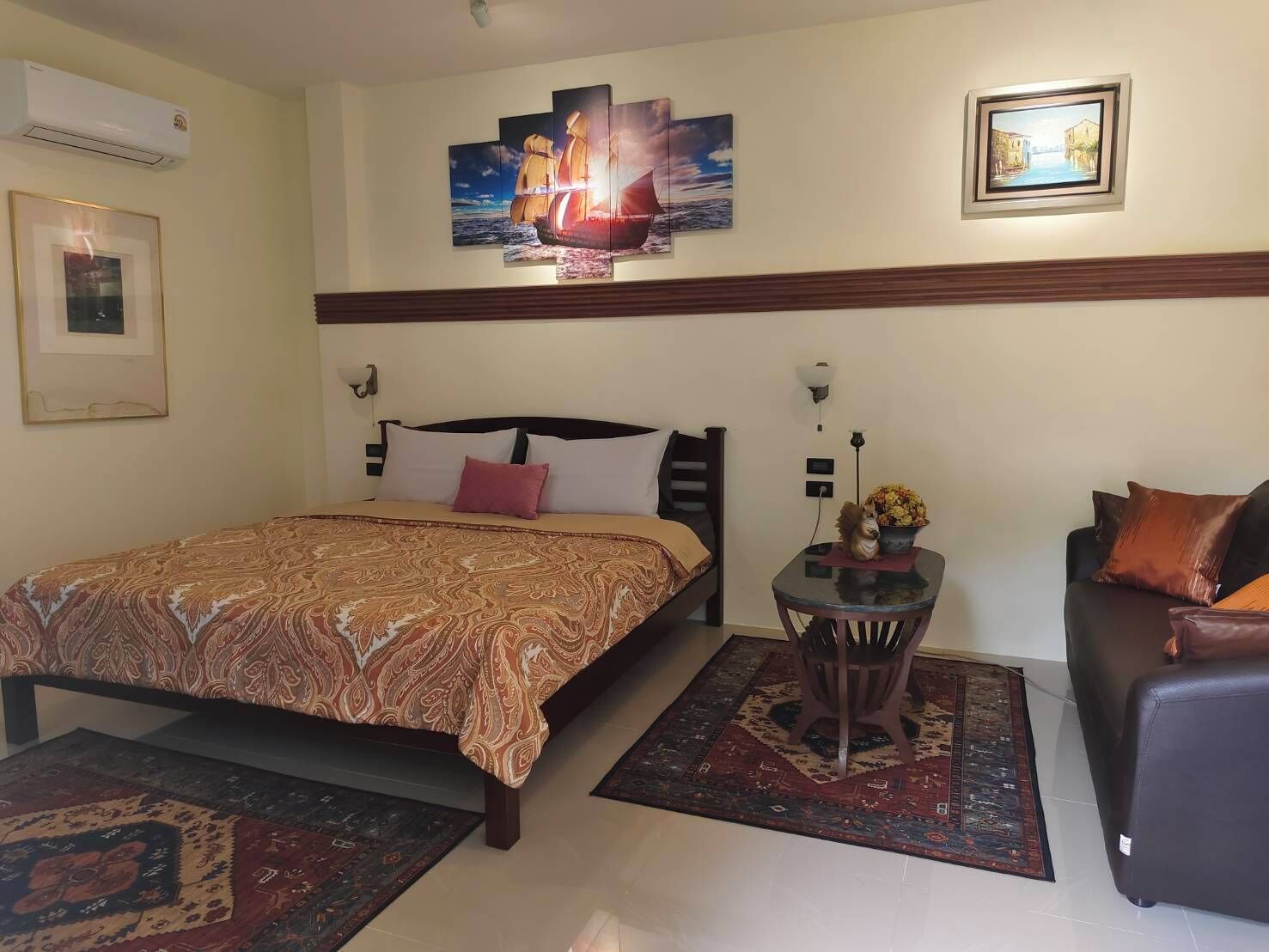 A hotel room with a bed, a sofa, a small table with a vase, and a painting on the wall.