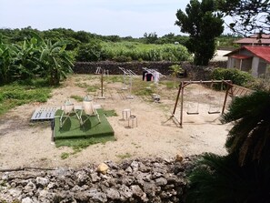 A rustic outdoor area with a dirt ground, a small garden with banana plants, a clothesline, and a swing set.