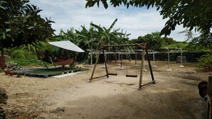 A playground with swings and a hammock, surrounded by lush greenery and a dirt ground. - Kohamaso (Taketomi)
