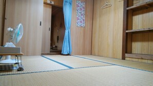 A room with tatami mat flooring, a standing fan, a bed with a white blanket, and a wooden wardrobe. - Kohamaso (Taketomi)