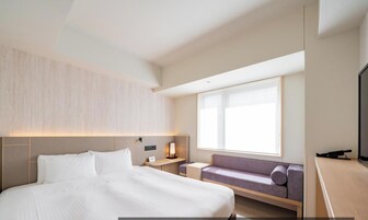 A modern hotel room with a large bed, a seating area, a TV, and a window providing natural light.