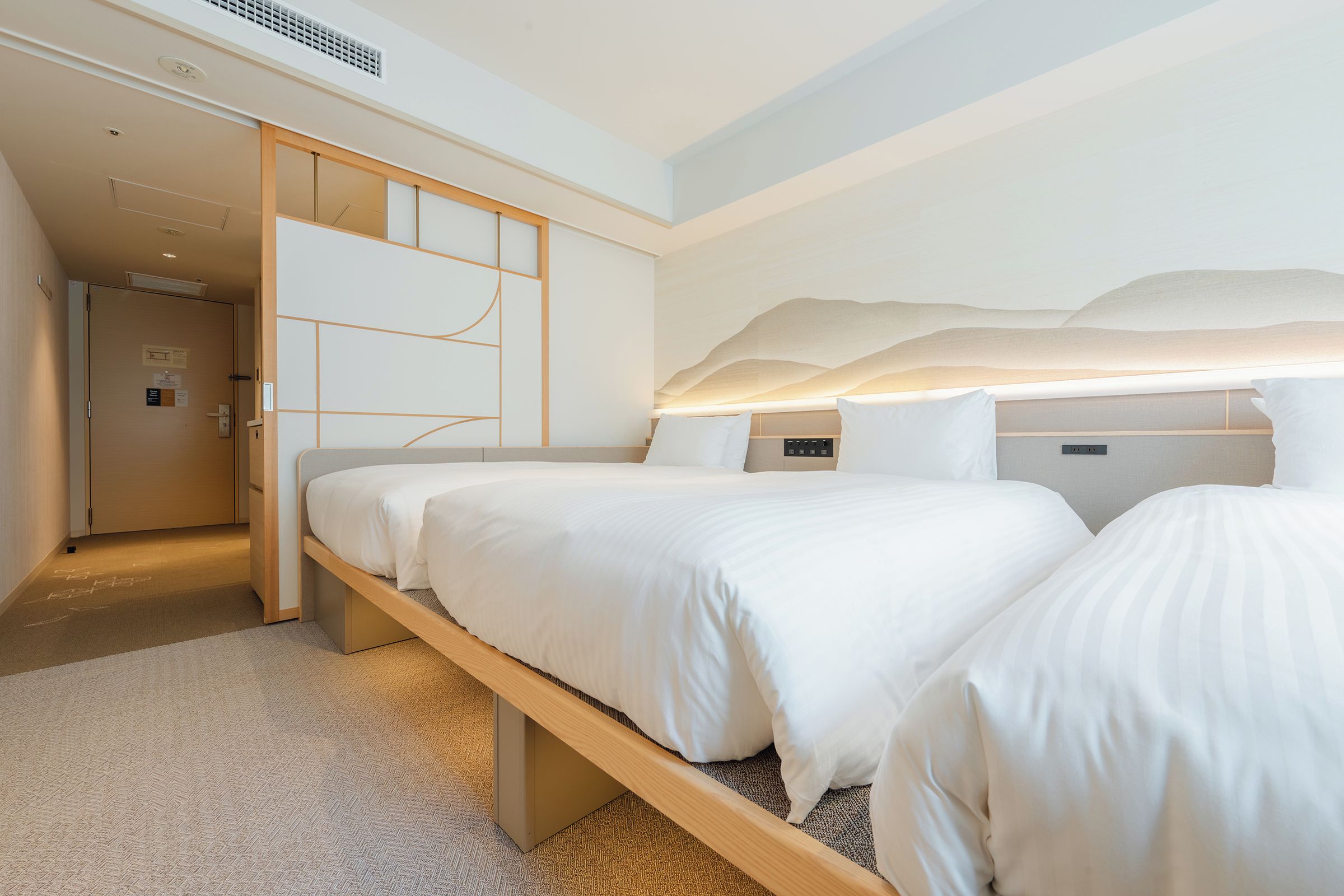 Ein Hotelzimmer mit einem Bett, einem Nachttisch, einer an der Wand montierten Steuerung und einer Tür zu einem anderen Raum.