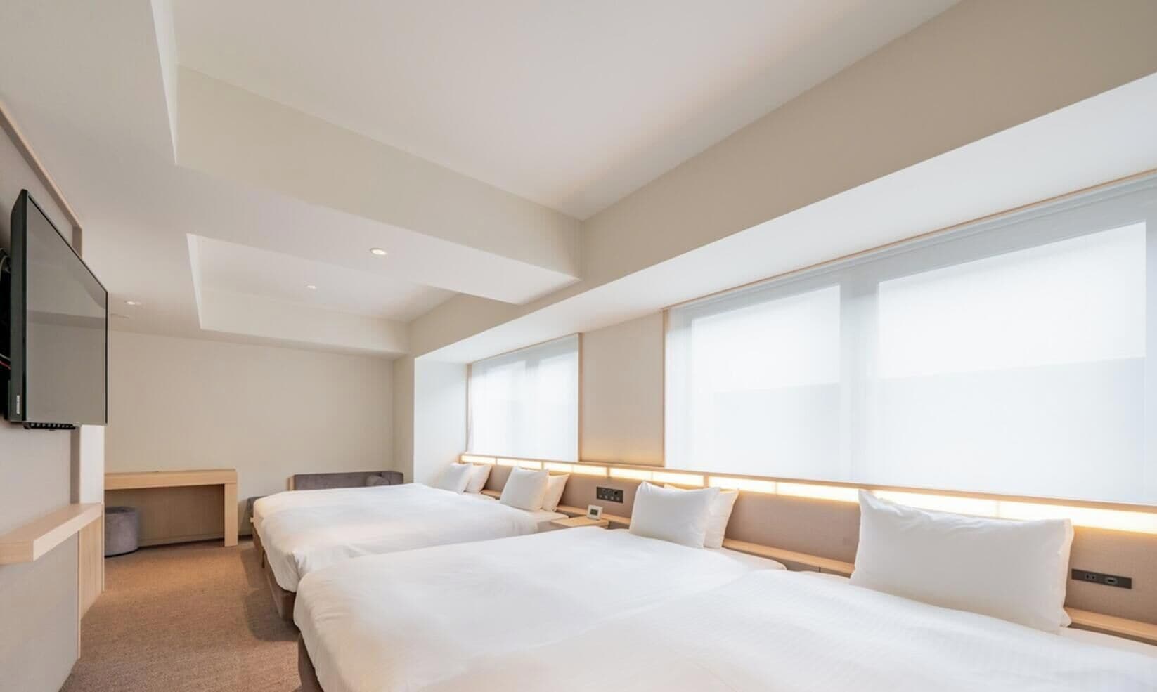 A hotel room with two beds, a desk, a TV, and large windows.