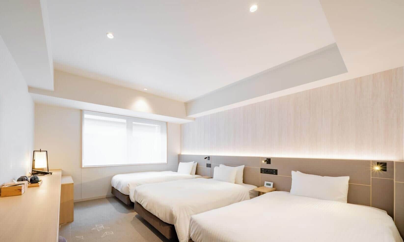 A hotel room with two beds, a desk, a window, and a wall-mounted light fixture.