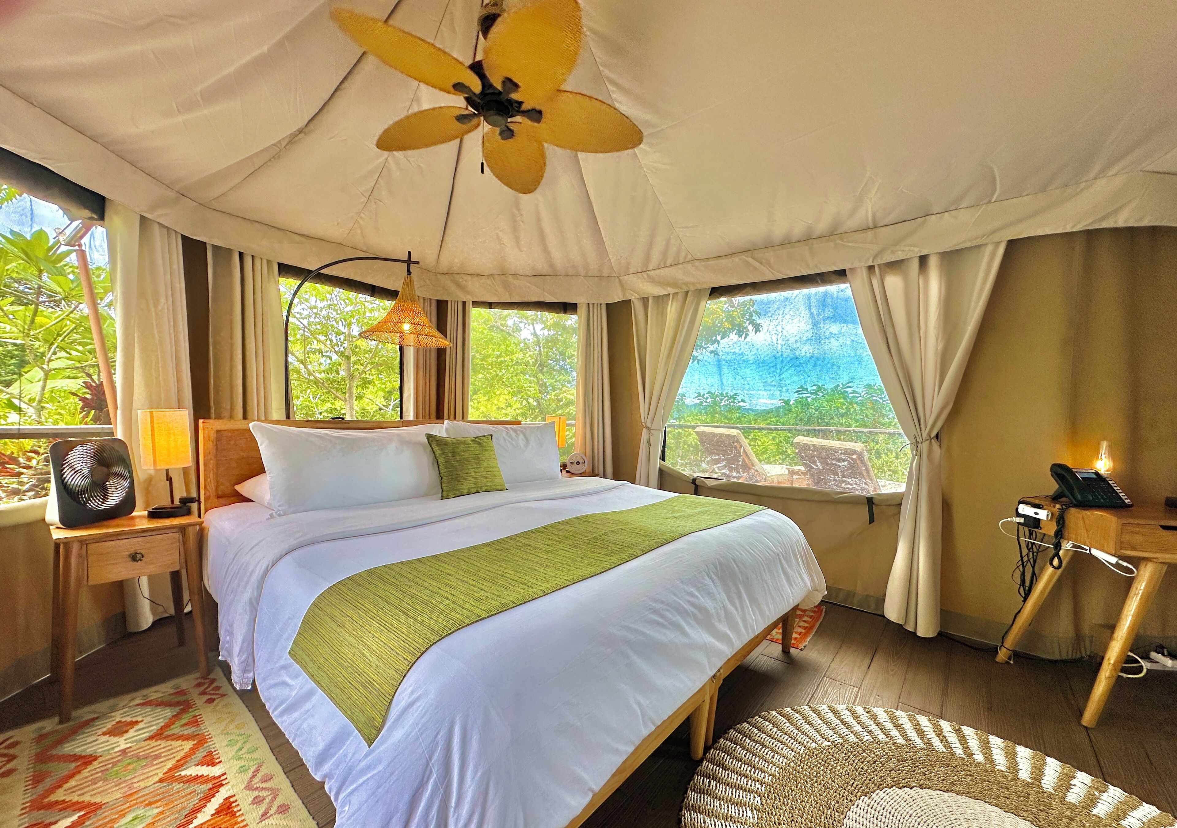 A bedroom with a bed, a ceiling fan, a bedside table with a lamp, a phone, and a view of the outdoors.