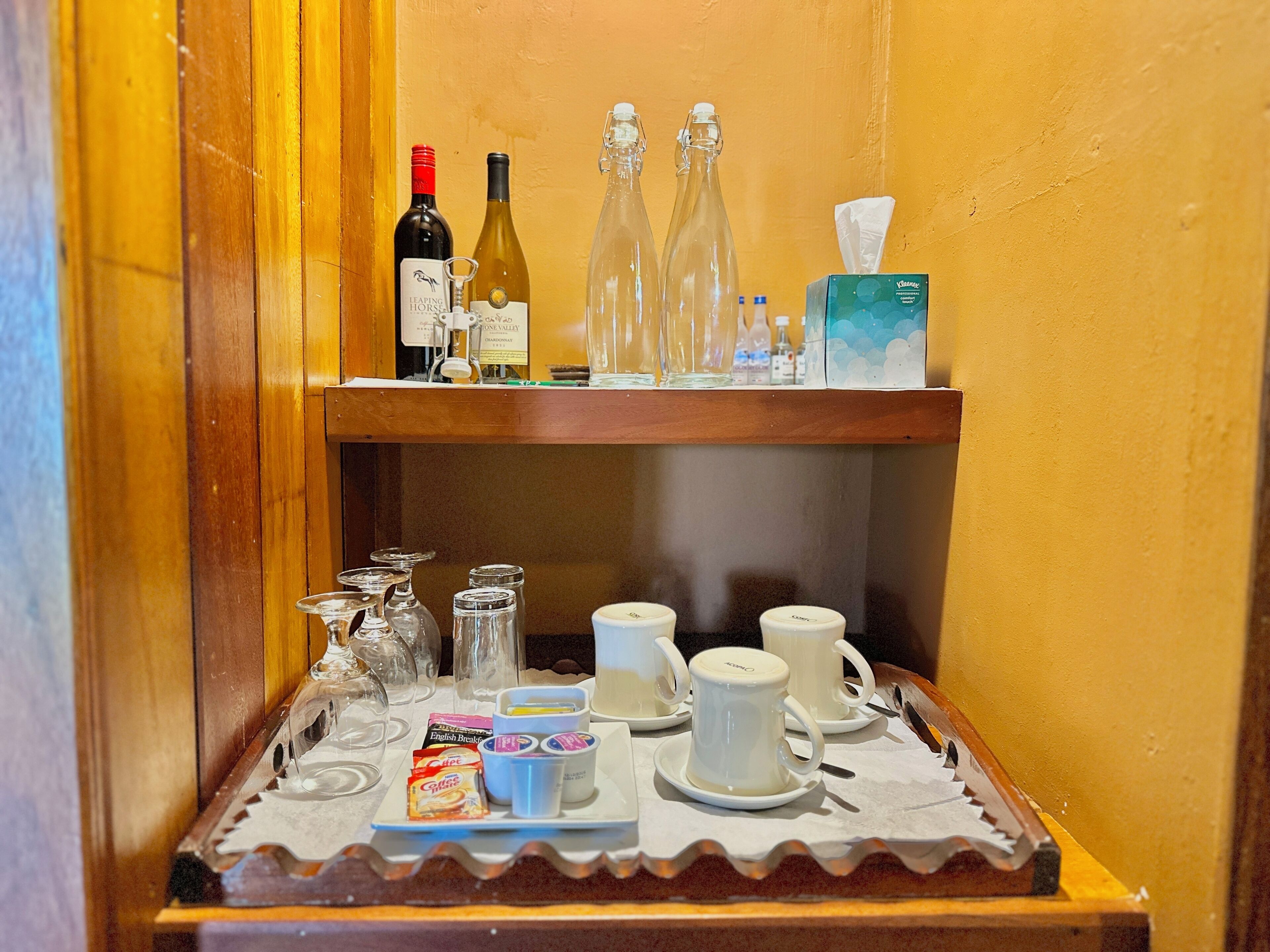 A bar area with a wooden shelf holding wine bottles, glasses, and a tissue box. Below, a tray with coffee packets, cups, and sugar cubes is visible.