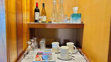 A bar area with a wooden shelf holding wine bottles, glasses, and a tissue box. Below, a tray with coffee packets, cups, and sugar cubes is visible.