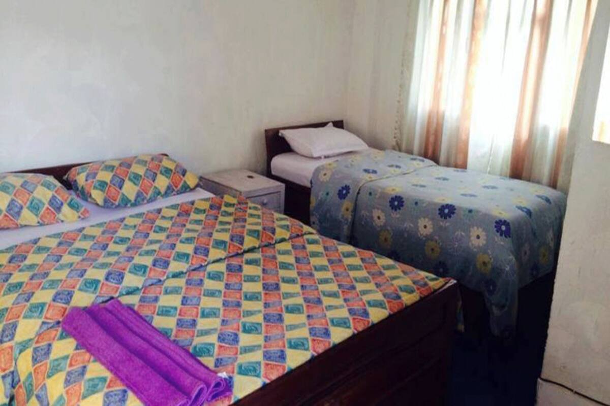 A room with two beds, one with a colorful checkered bedspread and the other with a floral one. There is a nightstand with a lamp between the beds.