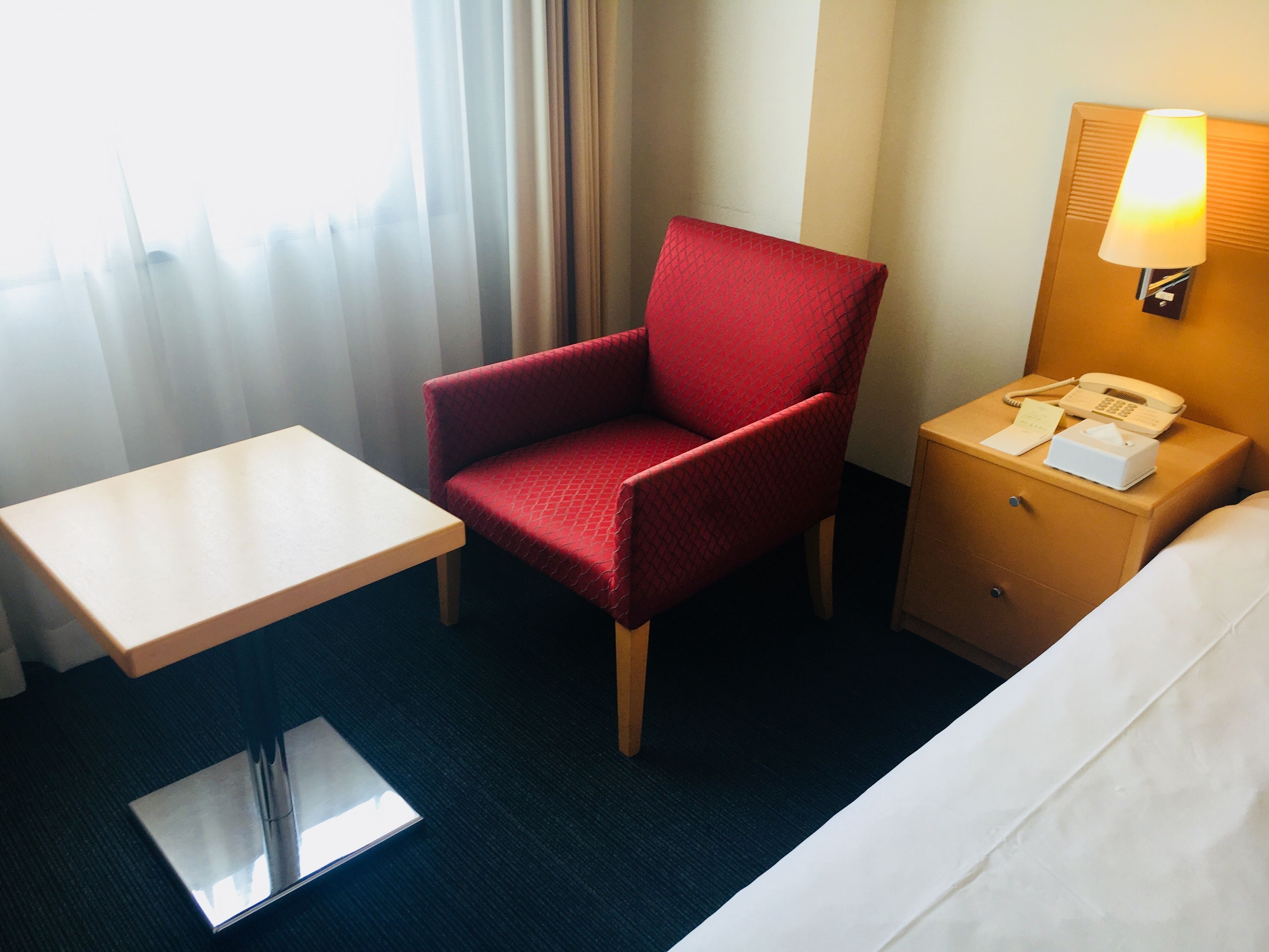 Habitación de hotel con un sillón rojo, una mesita, una mesita de noche con lámpara y una cama.