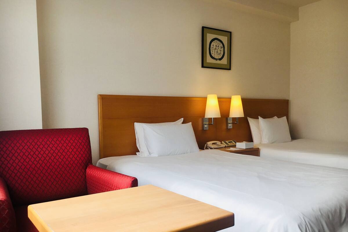 A hotel room with two beds, a red armchair, a wooden nightstand with a telephone, and two wall-mounted lamps.
