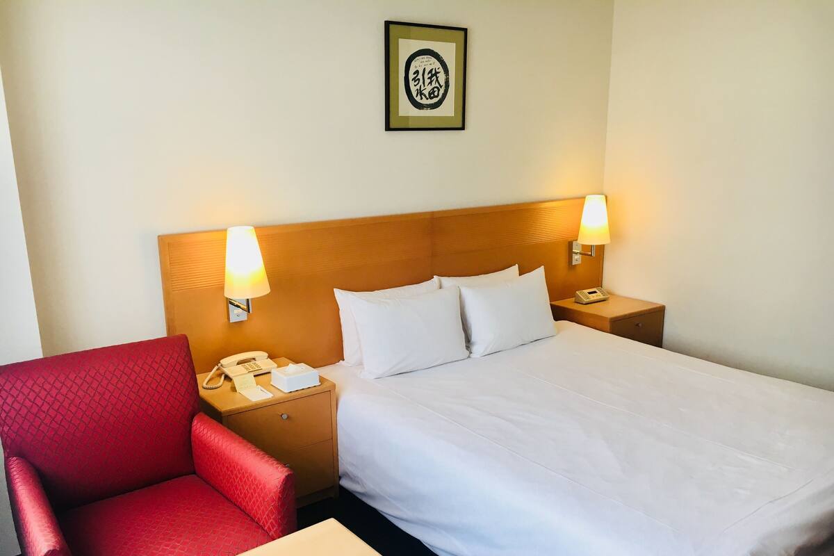 A hotel room with a bed, two bedside lamps, a telephone, a red armchair, and a small table.