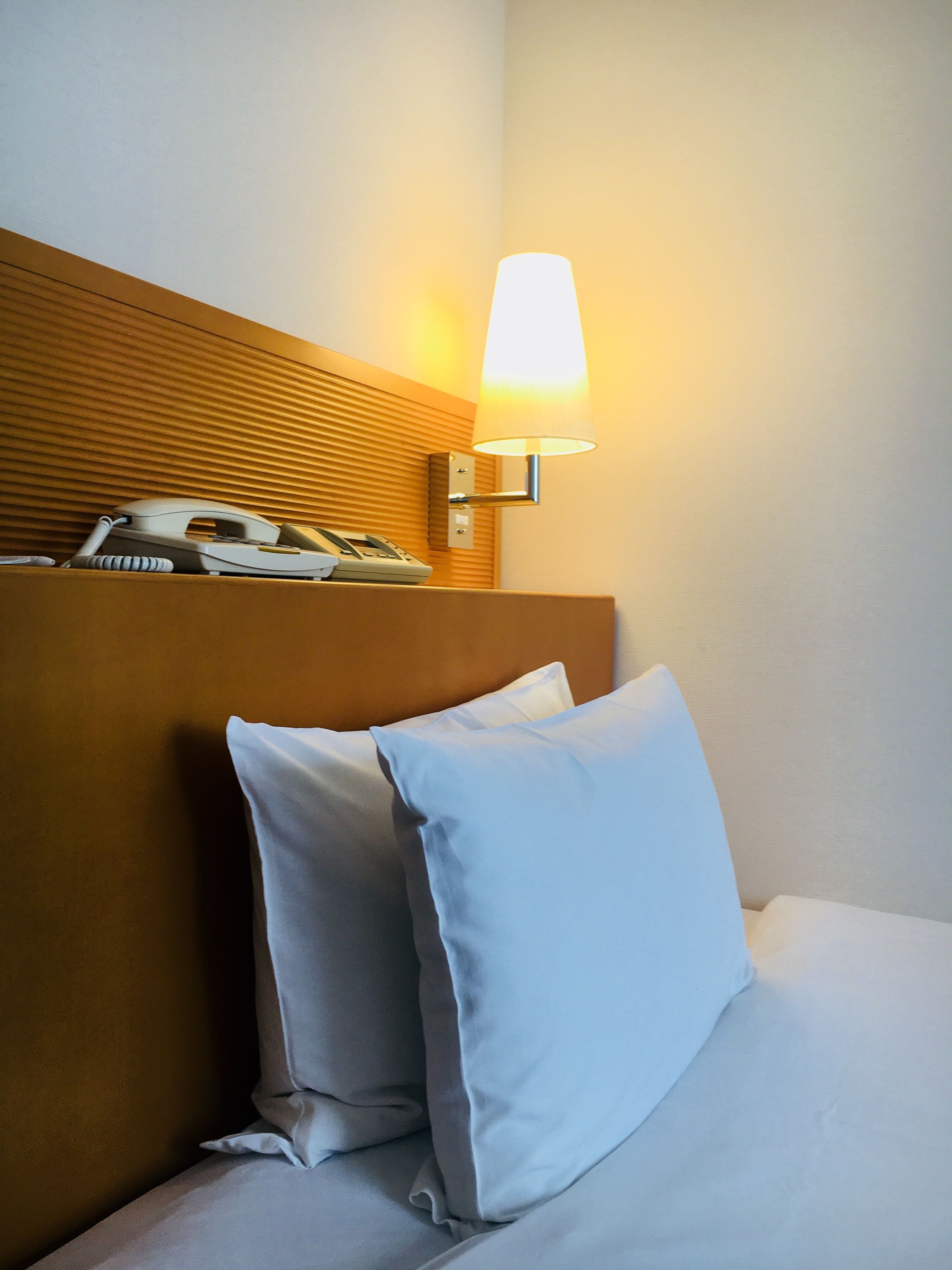 A hotel room with a bed, pillows, a bedside lamp, and a telephone on the headboard.