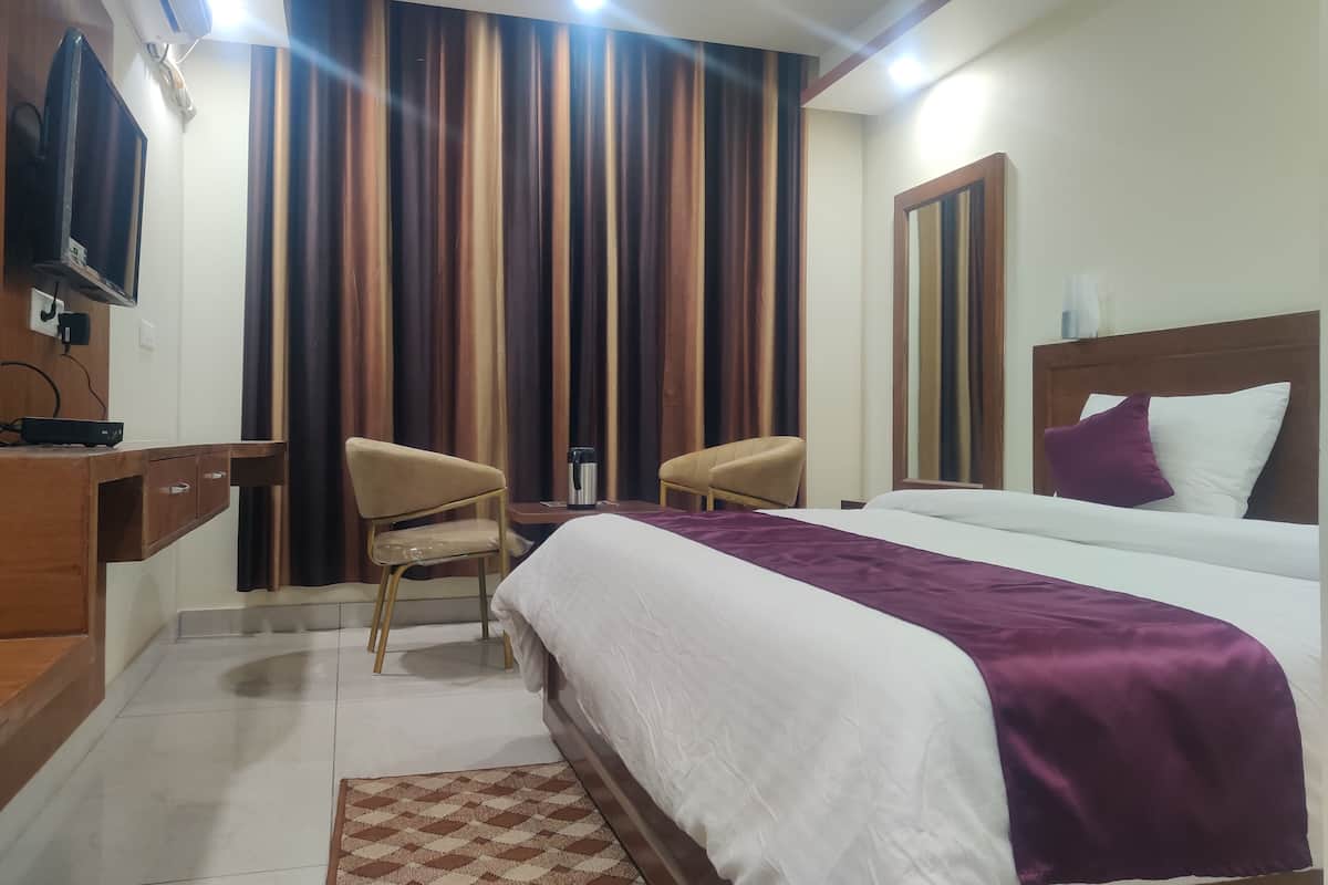A hotel room with a bed, two chairs, a desk with a TV, and a large window with curtains.