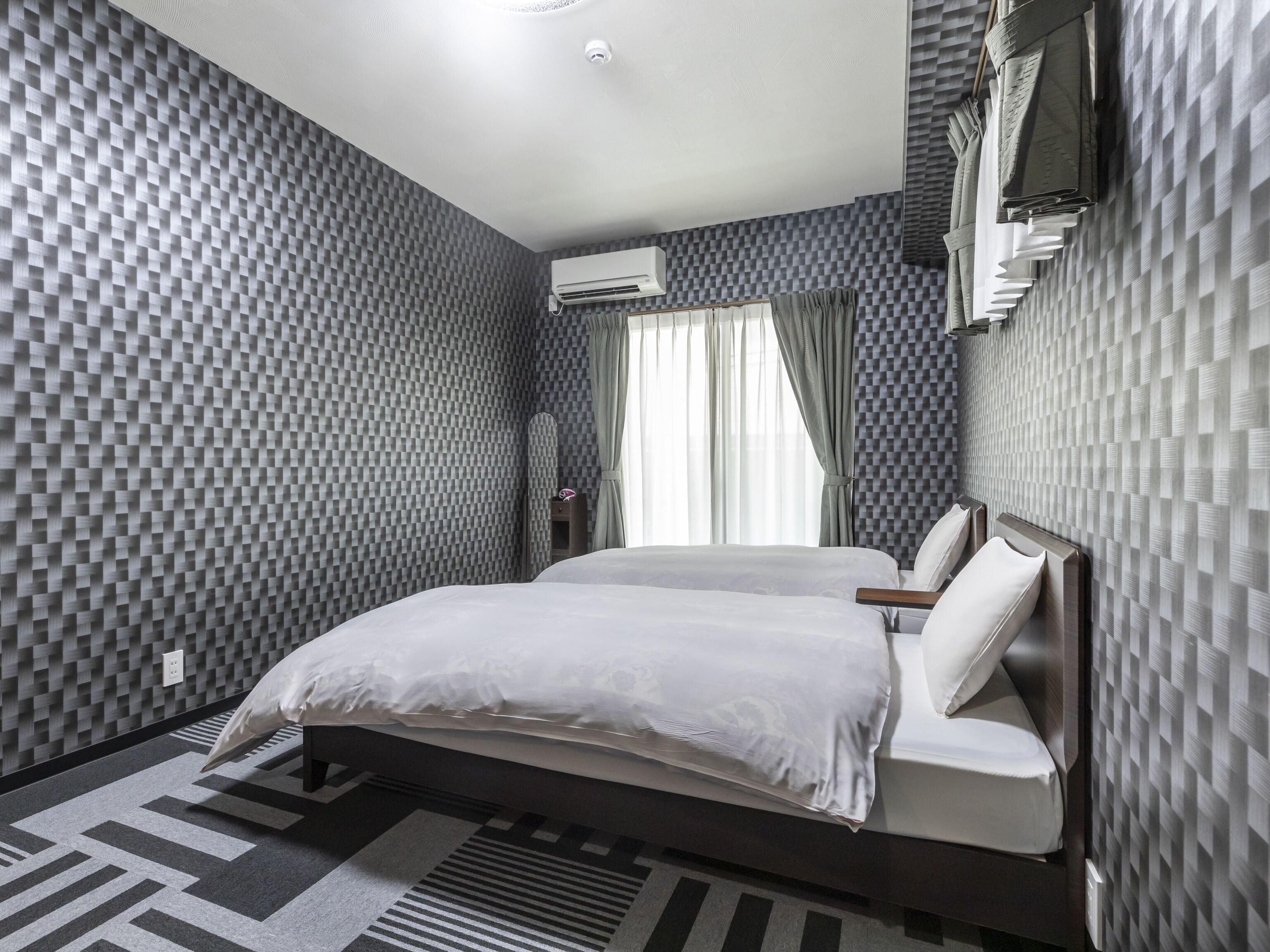 Ein modernes Hotelzimmer mit kariertem Wandmuster, einem Bett mit weißen Leinen und einer Klimaanlage an der Wand.