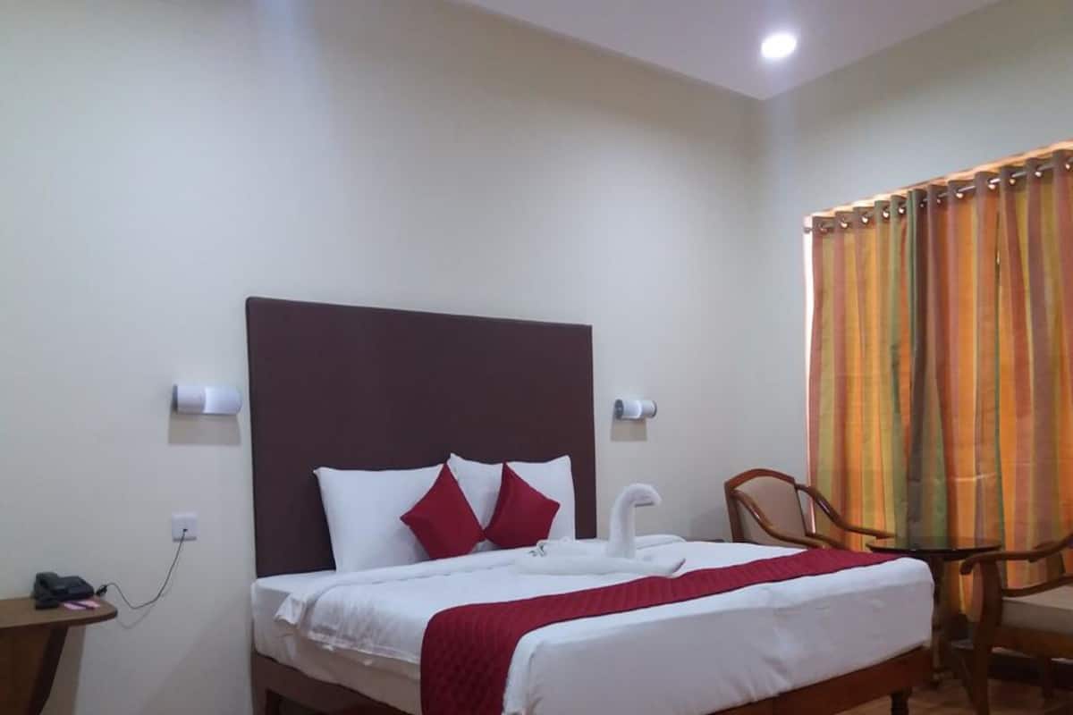 Una cama bien hecha con ropa de cama blanca y detalles en rojo, un cabecero de madera, una silla de madera y una mesita con un teléfono y cargador.