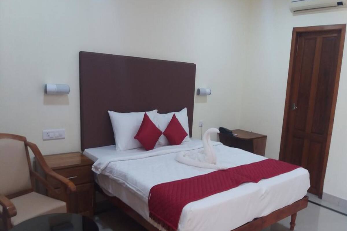 Habitación de hotel ordenada con una cama, una silla, una mesita pequeña y una mesita de noche de madera.