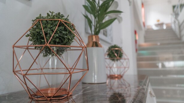 A hallway with a geometric copper wire plant stand, a white vase with a plant, and a staircase in the background. - Rinn Boutique House (Chiang Mai)