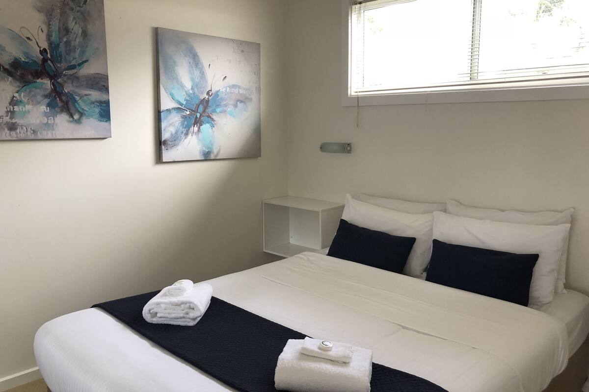 Una cama bien hecha con ropa de cama blanca y negra, un par de toallas dobladas y un cuadro abstracto en la pared.