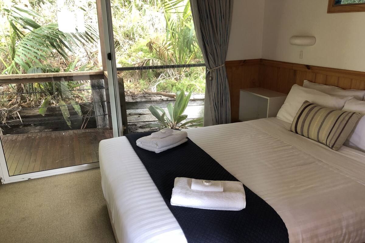 Habitación de hotel con cama, mesita de noche, ventana con cortinas y vista a zonas verdes.