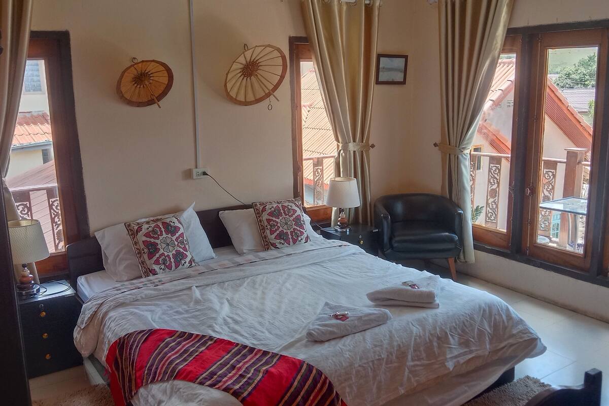 A bedroom with a bed, pillows, a chair, a nightstand, and a window with curtains.
