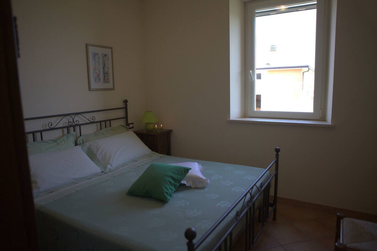 Een slaapkamer met een bed, een raam, een nachtkastje en een aan de muur hangend schilderij.