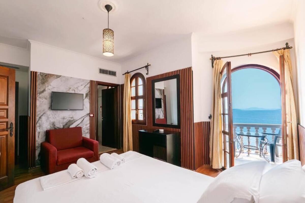 Habitación de hotel con cama, un sillón rojo, televisión y vista al mar a través de una ventana abierta.