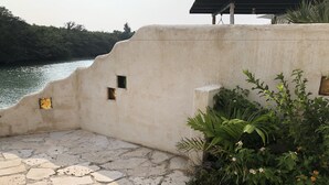A stone-tiled patio with a view of a body of water, a concrete wall with small windows, and lush greenery. - Lagoon Villa Ryusei (Miyakojima)