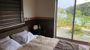 A bedroom with a bed, a nightstand, a vase, and a view of a lake and greenery. - Lagoon Villa Ryusei (Miyakojima)