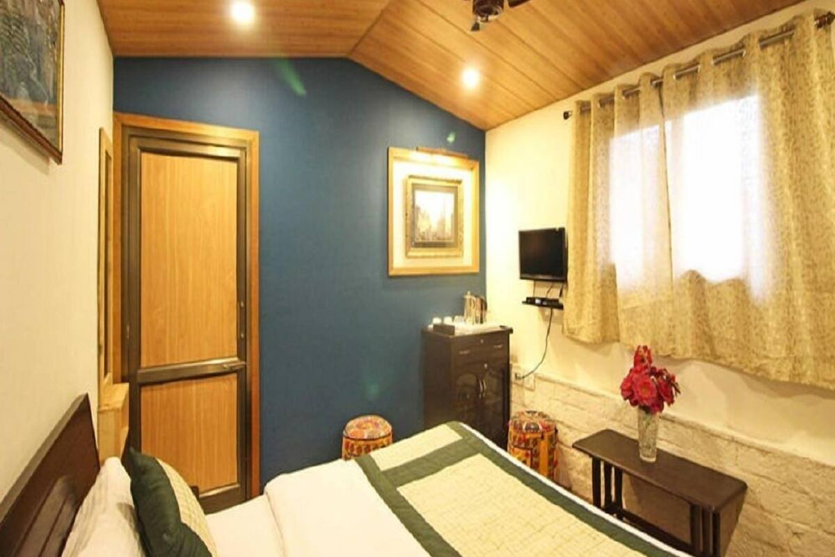Un dormitorio con techo de madera, una cama con colcha verde y blanca, un armario de madera, una mesita con un florero y un televisor en la pared.
