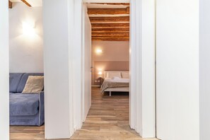 A narrow hallway leading to a bedroom with a bed and a nightstand, a blue sofa with a cushion, and a wooden ceiling.