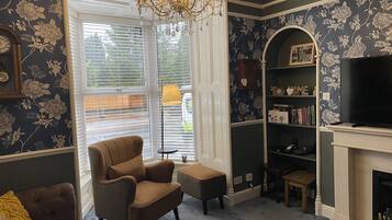 A living room with a dark floral wallpaper, a brown armchair, a bookshelf, a fireplace, and a chandelier.