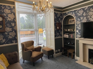 A living room with a dark floral wallpaper, a brown armchair, a bookshelf, a fireplace, and a chandelier.